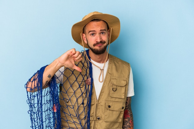 Jeune pêcheur caucasien avec des tatouages tenant un filet isolé sur fond bleu montrant un geste d'aversion, les pouces vers le bas. Notion de désaccord.