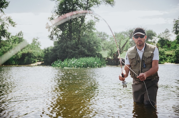 Jeune pêcheur pêchant sur lac ou rivière. Un gars sérieux et concentré en vêtements de pêcheur se tient dans l'eau de la rivière ou du lac et tient la tige. Essayer d'attraper de délicieux poissons savoureux. Passe-temps ou style de vie.