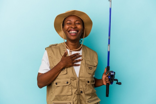 Jeune pêcheuse afro-américaine tenant une tige isolée sur fond bleu rit fort en gardant la main sur la poitrine.