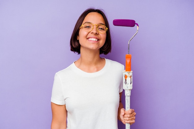 Jeune peintre métisse femme tenant un bâton de peinture isolé sur fond violet heureux, souriant et joyeux.