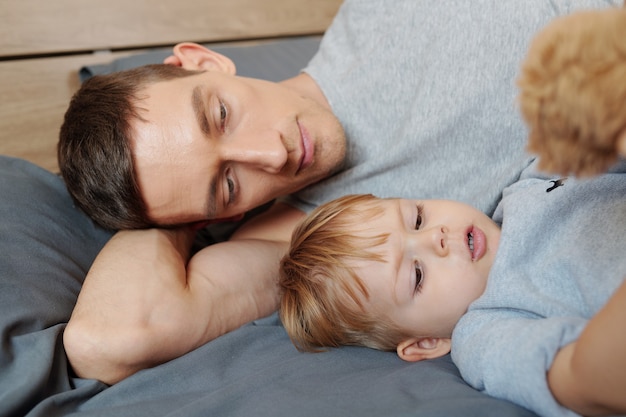 Jeune père allongé sur le lit et se reposant avec son petit fils pendant l'heure du sommeil