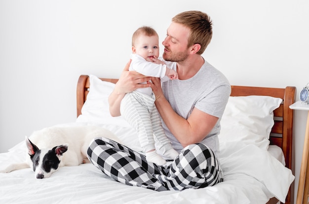 Jeune père assis dans le lit avec un petit fils et un chien mignon dans la pièce avec la lumière du soleil et serrant son petit garçon dans ses bras. Homme restant dans la chambre avec son enfant et son animal de compagnie. Mec avec enfant et chien le matin