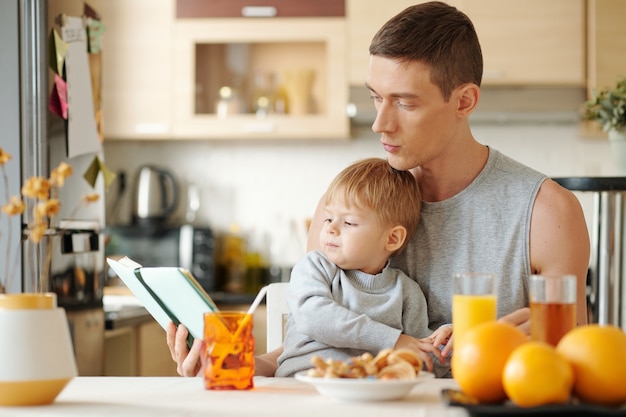 Jeune père assis à table avec son petit fils et lui lisant un livre pendant le petit-déjeuner