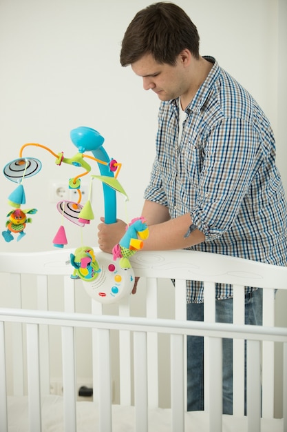 Jeune père attentionné assemblant le lit de bébé et y mettant un carrousel de jouets