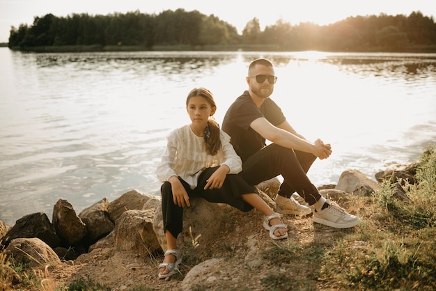 Un jeune père avec une barbe et des lunettes de soleil pose sur des pierres avec sa fille sur la côte du lac le soir. Famille monoparentale élégante en vacances.