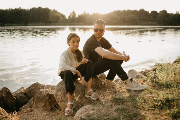 Un jeune père avec une barbe et des lunettes de soleil pose sur des pierres avec sa jolie fille au bord du lac. Famille monoparentale élégante en vacances au coucher du soleil.