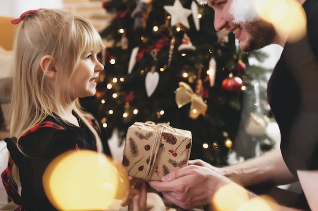 Jeune père en bonnet rouge donnant un cadeau de Noël à sa fille souriante près de l'arbre de Noël décoré. Fille vêtue d'une tenue festive de Noël rouge-noir. Ils sourient, heureux à cause du Nouvel An