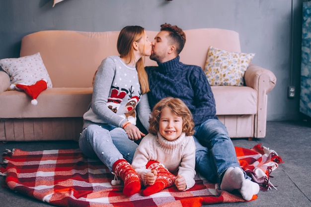 Jeune père de famille, mère et fils vêtus de chandails de Noël sont assis près du canapé dans une pièce confortable. Le fils allongé entre les parents regarde dans le cadre et sourit, dans le baiser des parents.