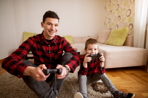 Jeune père et fils excités joyeux dans la même chemise rouge jouant à des jeux de console avec des manettes dans un salon lumineux.