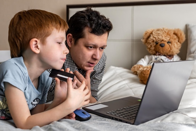 Jeune père et fils shopping en ligne ensemble temps en famille Enfant garçon aux cheveux roux et papa à l'ordinateur portable choisissent les achats Le garçon tient une carte de débit Paiement Internet