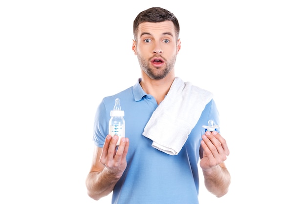 Jeune père frustré. Jeune homme choqué avec une serviette sur l'épaule tenant une bouteille avec du lait et une tétine en se tenant debout sur fond blanc