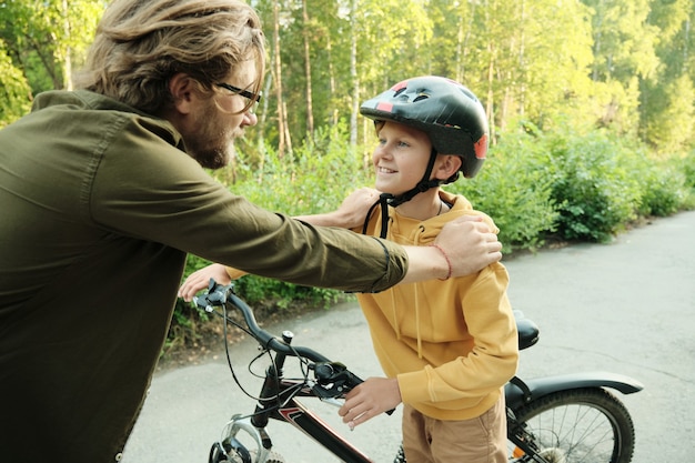 Jeune père mettant les mains sur les épaules de son jeune fils heureux tout en lui donnant des instructions avant de faire du vélo dans le parc le week-end