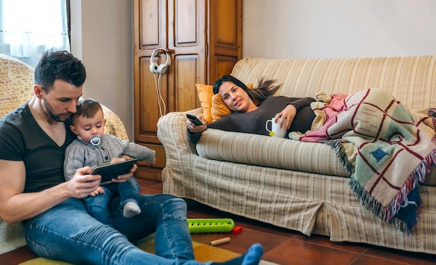 Jeune père regardant la tablette avec son petit fils pendant que la mère enceinte regarde la télévision