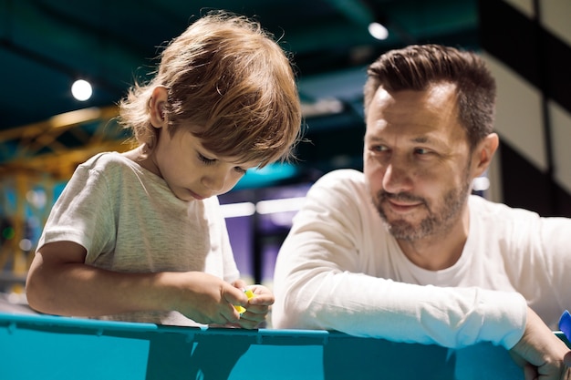 Le Jeune Père Regarde Son Fils Jouer Avec Tendresse, Ils Passent Du Temps Ensemble Dans Un Centre De Développement. Amour Et Soutien Parental. Centres De Développement Pour Enfants.