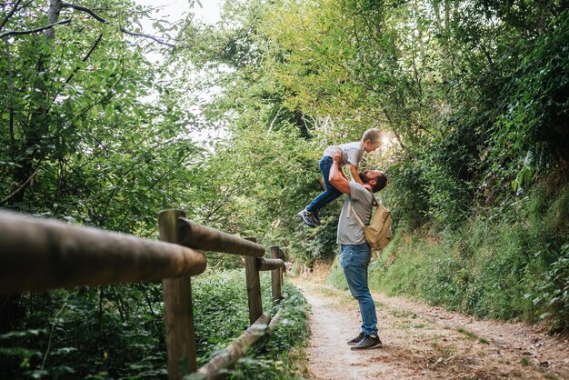 Photo jeune père avec sac à dos tenant un garçon de 6 ans dans la forêt pendant qu'ils rient