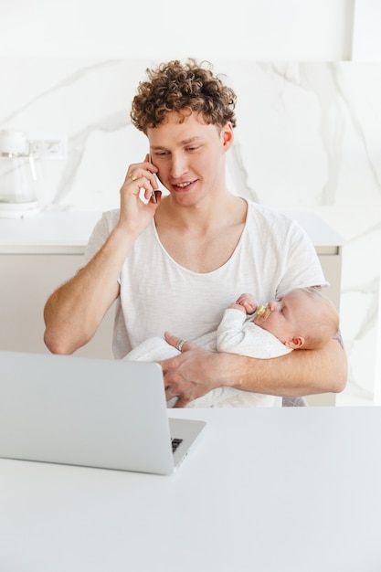 Jeune Père Séduisant Travaillant Sur Un Ordinateur Portable Assis à La Table à La Maison Et Tenant Son Bébé