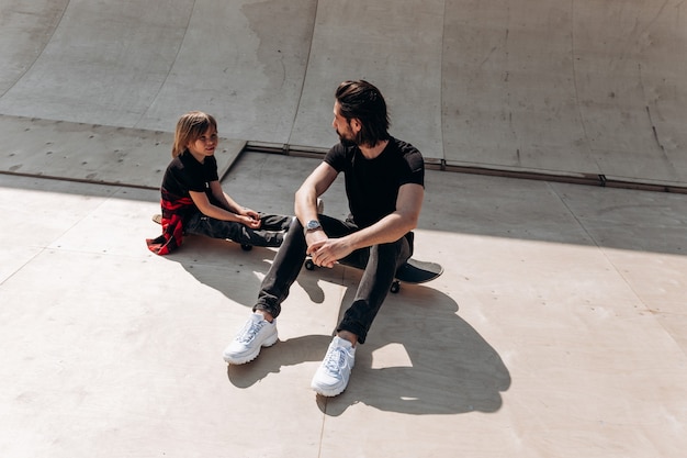 Le jeune père et son fils vêtus de vêtements décontractés sont assis sur les planches à roulettes dans un skate park à la journée ensoleillée.