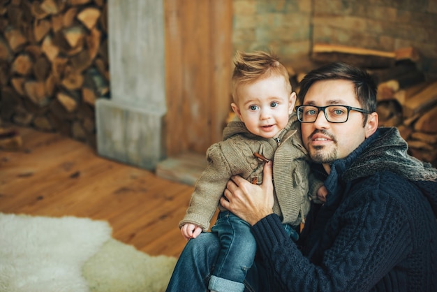 Jeune père et son petit fils portant des vestes tricotées posant près de la cheminée. Réveillon de Nouvel an. Vacances confortables, amour, concept de famille heureuse.