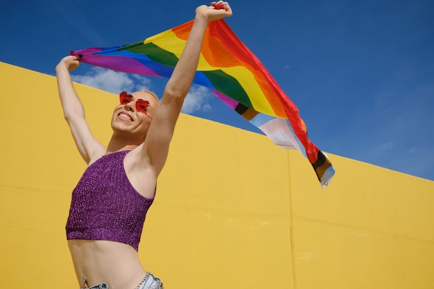 Jeune personne non binaire ayant l'air heureuse et excitée tout en agitant un drapeau arc-en-ciel à l'extérieur. Concept d'égalité, de droits et d'identité de genre.