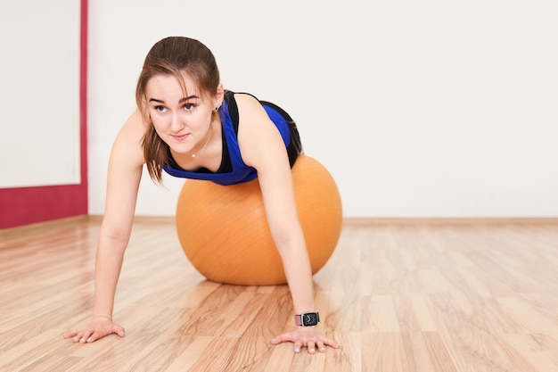 Jeune petite femme s'entraîne en faisant des pompes avec un ballon d'exercice dans une salle de sport