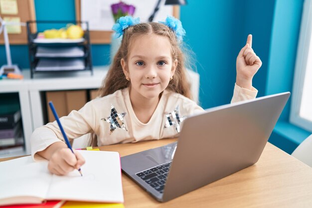 Jeune petite fille assise sur la table à faire ses devoirs avec un ordinateur portable souriant heureux pointant avec la main et le doigt sur le côté