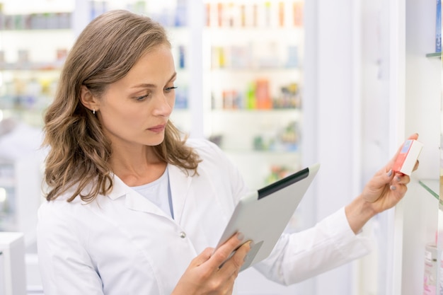 Jeune pharmacien avec contrôle du pavé tactile ou à la recherche de nouveaux médicaments dans la base de l'ordinateur en se tenant debout par l'un des affichages en pharmacie