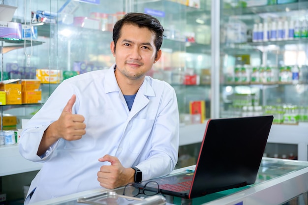 Jeune pharmacien de sexe masculin souriant joyeusement dans la pharmacie faisant l'inventaire dans une pharmacie bien approvisionnée et à jour. Pharmacien utilisant l'ordinateur à la pharmacie