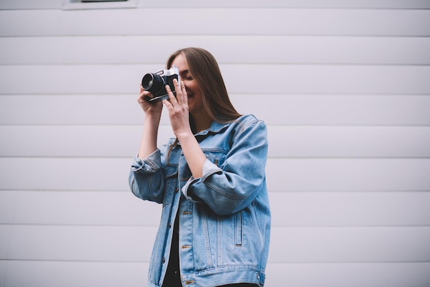 Jeune photographe amateur souriante faisant des photos à l'aide d'un appareil photo vintage près du mur à l'extérieur