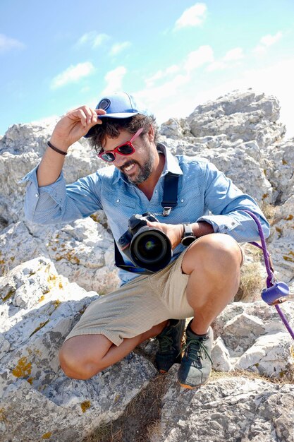 Jeune photographe aventureux au bord de la falaise souriant en bonnet bleu et vêtements bleus.