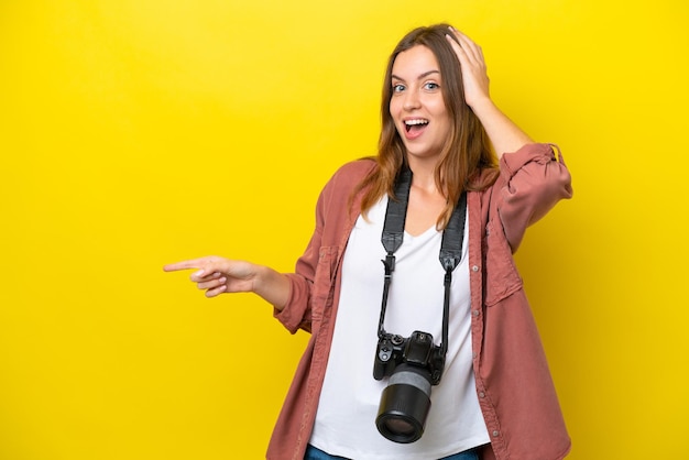 Jeune photographe caucasien femme isolée sur fond jaune surpris et pointant le doigt sur le côté