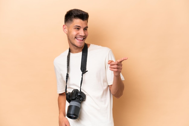 Jeune photographe caucasien homme isolé sur fond beige pointant le doigt sur le côté et présentant un produit