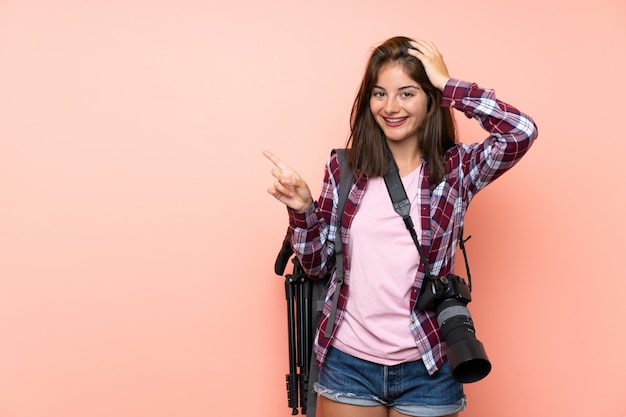 Jeune photographe fille sur mur rose isolé surpris et pointant le doigt sur le côté