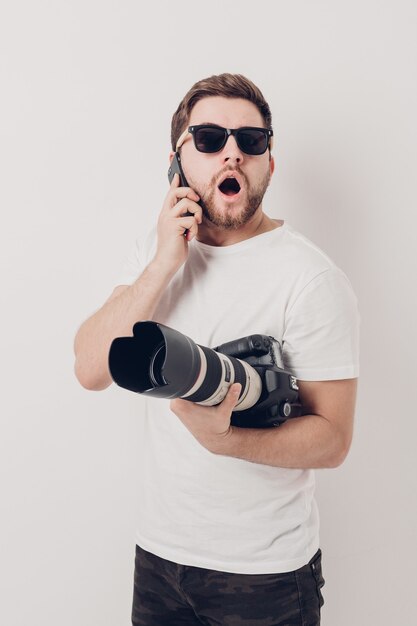 Un jeune photographe professionnel en chemise blanche tient un appareil photo numérique lourd avec un objectif long et parle au téléphone avec un client