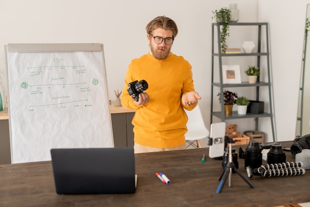 Jeune photographe professionnel en tenue décontractée en regardant la caméra du smartphone et l'affichage de l'ordinateur portable tout en parlant au public en ligne