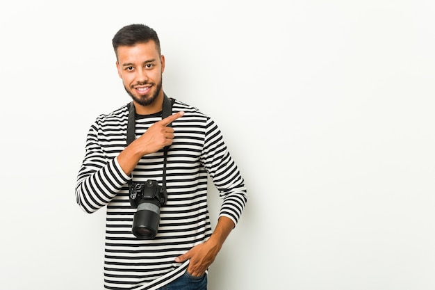 Jeune photographe sud-asiatique souriant et pointant de côté, montrant quelque chose à l'espace vide.