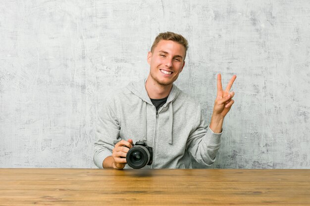 Jeune photographe tenant une caméra sur une table joyeuse et insouciante montrant un symbole de la paix avec les doigts.