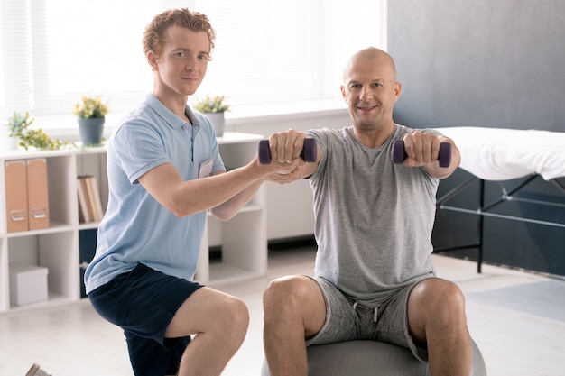 Jeune physiothérapeute aidant un patient masculin sur fitball à faire de l'exercice avec des haltères tout en soutenant son bras droit dans des cliniques de rééducation