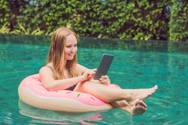 Jeune pigiste assise près de la piscine avec son ordinateur portable à l'hôtel naviguant dans son smartphone. Occupé pendant les vacances. Concept de travail à distance. Copiez l'espace pour votre texte