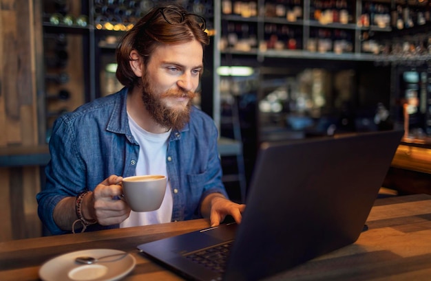 Jeune pigiste barbu vérifiant ses e-mails via un ordinateur portable tout en prenant un café le matin dans un bar loft