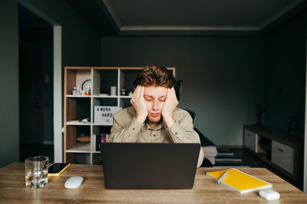 Jeune pigiste fatigué travaillant à la maison sur un ordinateur portable au visage triste et s'endormant Le télétravailleur fait un travail ennuyeux Portrait d'un pigiste endormi et fatigué Quarantaine