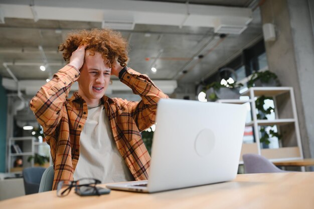 Photo jeune pigiste masculin heureux dans des vêtements décontractés assis dans un café avec un ordinateur portable et utilisant un téléphone portable