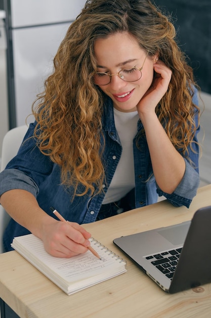 Jeune pigiste en tenue décontractée assise à table avec netbook et prenant des notes dans le bloc-notes tout en travaillant sur un projet