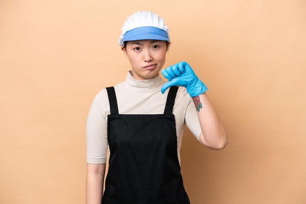 Jeune poissonnière chinoise portant un tablier et tenant un poisson cru isolé sur fond rose montrant le pouce vers le bas avec une expression négative