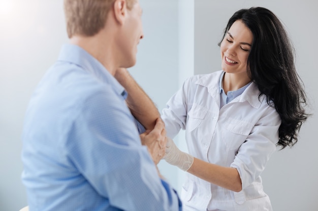 Jeune praticien qualifié positif travaillant à l'hôpital et aidant un patient mature tout en examinant le coude du patient