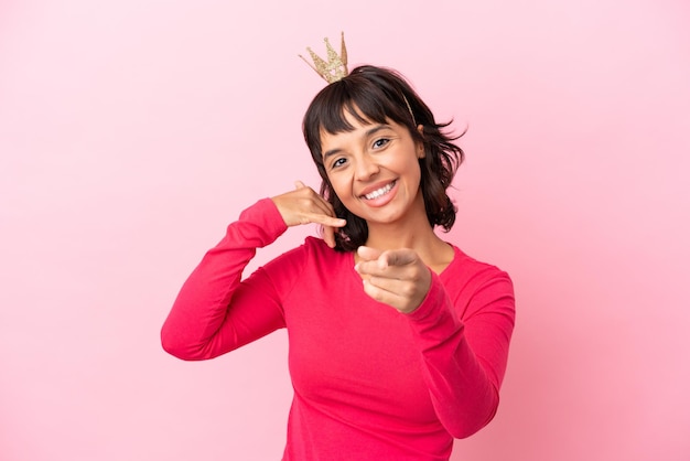Jeune princesse métisse avec couronne isolée sur fond rose faisant un geste de téléphone et pointant vers l'avant
