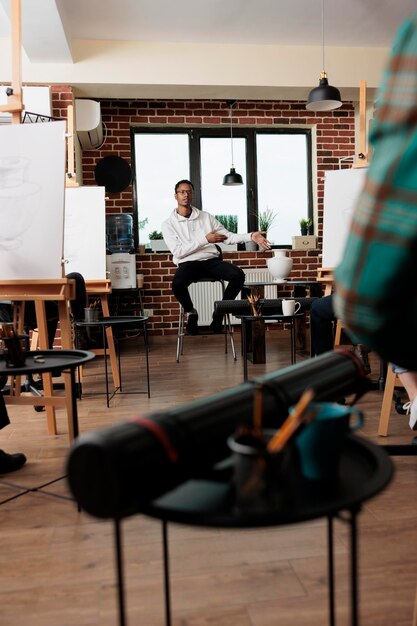 Photo jeune professeur afro-américain assis en classe à partir d'étudiants parlant des fondements du dessin de nature morte pendant le cours d'art de groupe prooffesion artiste masculin enseignant une leçon de dessin