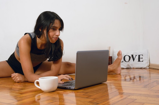 Une jeune professeur de yoga latine avec les jambes ouvertes assise sur le sol avec des cours de planification d'ordinateur portable