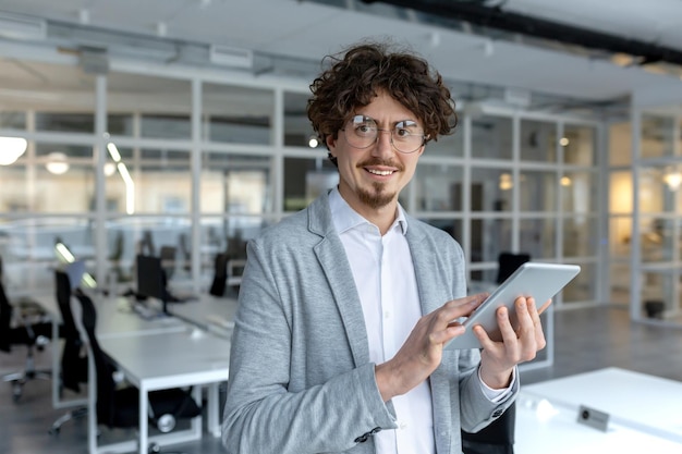 Un jeune professionnel souriant aux cheveux bouclés utilisant une tablette numérique dans un bureau contemporain lumineux