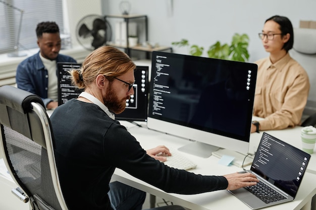 Jeune programmeur de diversité sérieux tapant sur un clavier d'ordinateur portable assis devant des ordinateurs par lieu de travail contre deux collègues