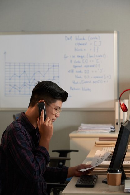 Jeune programmeur regardant un document avec un organigramme de projet et parlant au téléphone avec des collègues de...